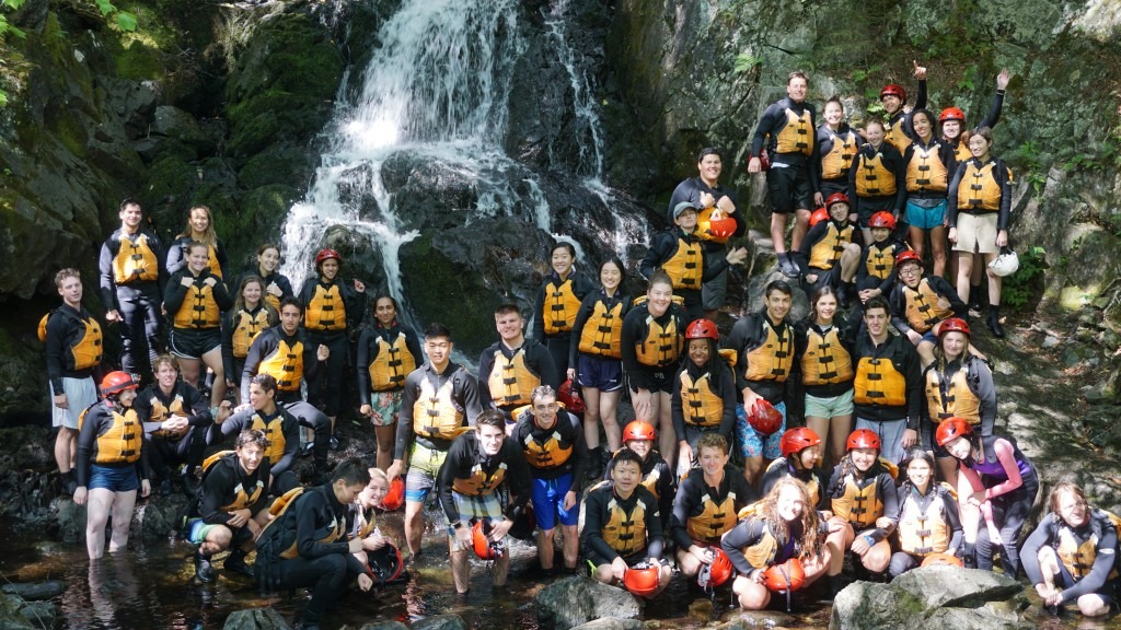 School group in rafting gear posing near a waterfall.